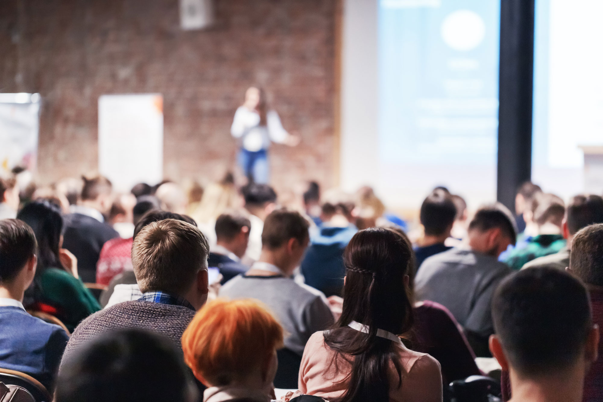A large group of people listening to a female speaker deliver a speech from a stage within an industrial office environment