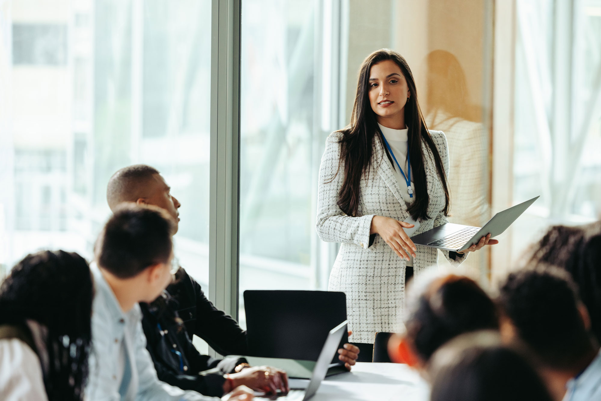A female leader stood up explaining something on her laptop to a group of colleagues sat down on a board table
